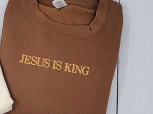 Brown sweatshirt folded on a white surface, text reads "JESUS IS KING" in yellow, conveying a spiritual message.