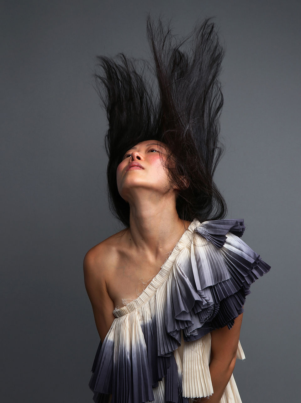"Fine art portrait by Christina Diebel: Asian woman with flying black hair in pleated gradient dress, captured in a moment of movement and devotion."