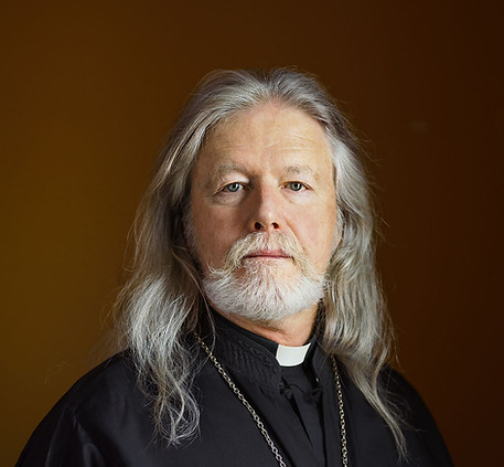 Father Stephan, a priest with long white hair, mustache and short beard, wearing a black clergy shirt with white collar and cross on chains
