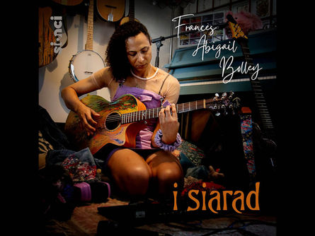 Music cover artwork with a photograph of a woman in a purple top playing an acoustic guitar with multiple musical instruments in the background.