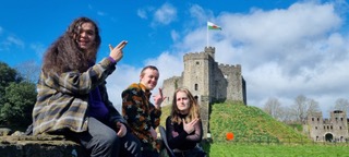 Three people sitting outside Cardiff Castle, gesturing at the camera with a cloudy blue sky in the background.