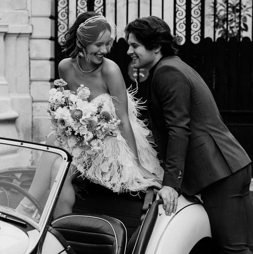 A couple smiles beside a vintage car. The woman, in a feathered dress, holds a bouquet. Elegant, joyful mood; wrought iron gate behind.