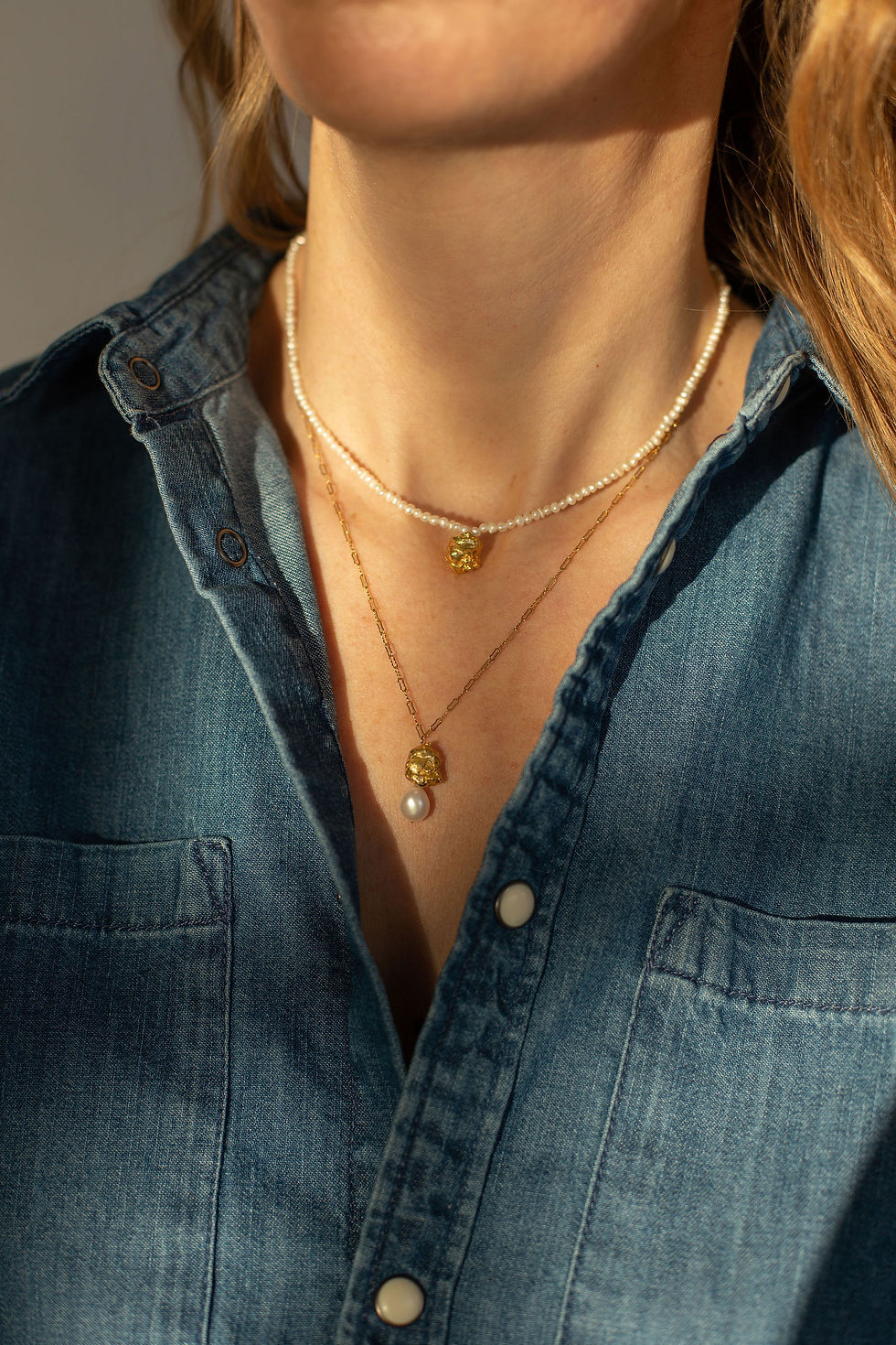 Close-up of a woman wearing two necklaces, one pearl and one gold with a charm. She's in a denim shirt, bathed in warm light.