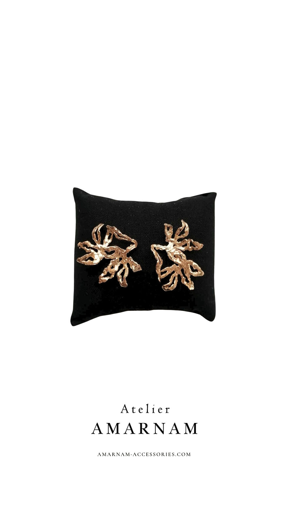 Gold leaf-shaped earrings on a black cushion. White background with "Atelier AMARNAM" and website below. Elegant and minimalist.