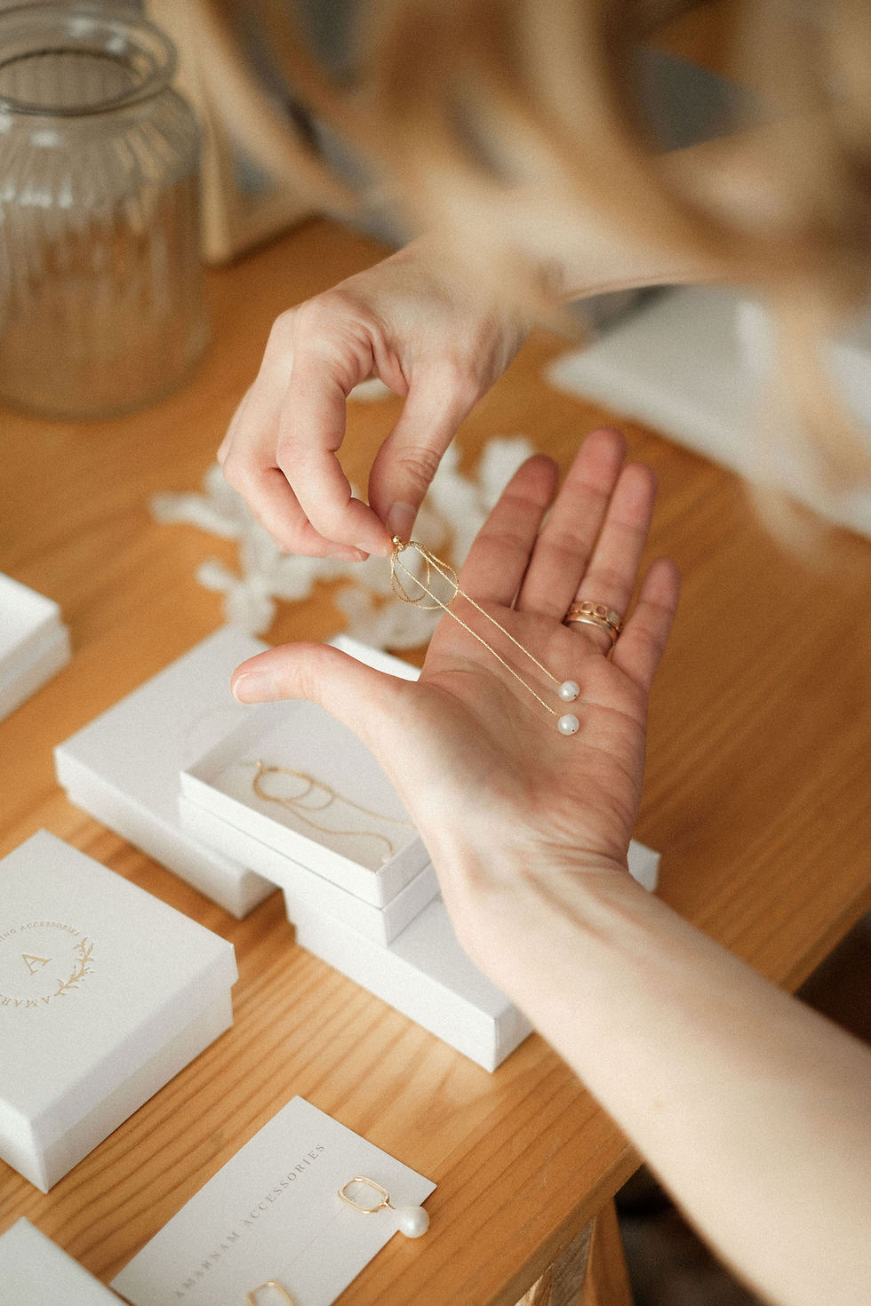 Hands holding a pearl earrings over a wooden table with white jewelry boxes and cards. Soft lighting creates an elegant, calm mood.