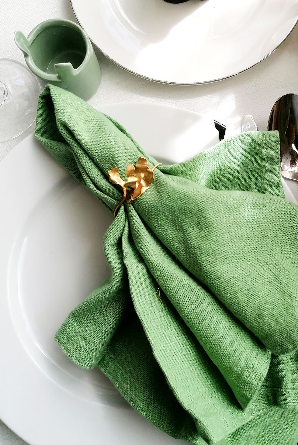 Green napkin with gold leaf holder on a white plate, beside a matching green creamer, set on a white tablecloth.