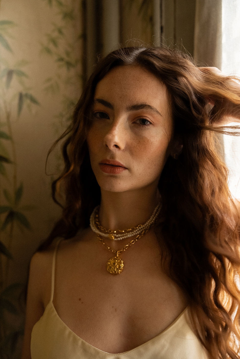 Woman with long red hair and golden necklaces stands by a window, wearing a cream dress. Green leaf patterned wallpaper in background.