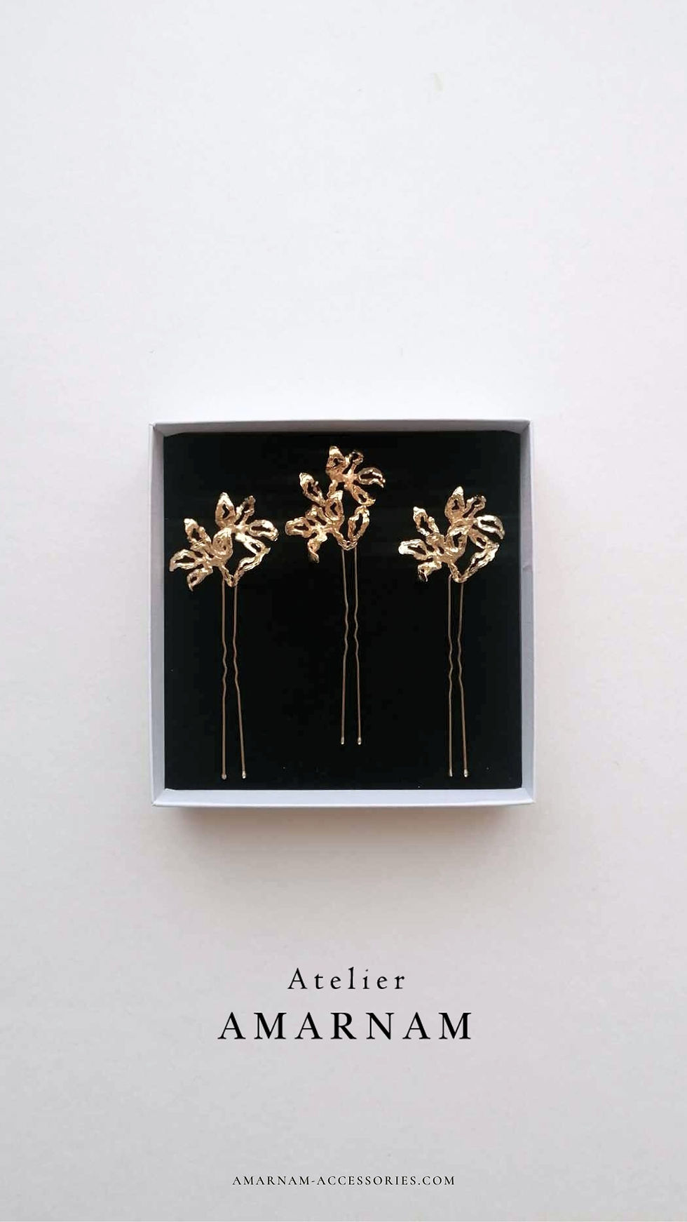 Gold floral hairpins in black box with "Atelier AMARNAM" text below. Simple white background, elegant and minimal design.