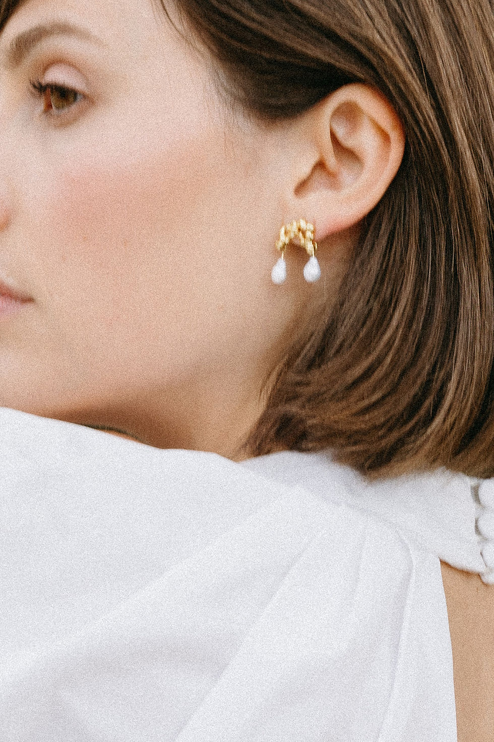 Woman in white top wearing gold earrings with white drops, looking thoughtful. Brown hair, neutral background, close-up profile view.
