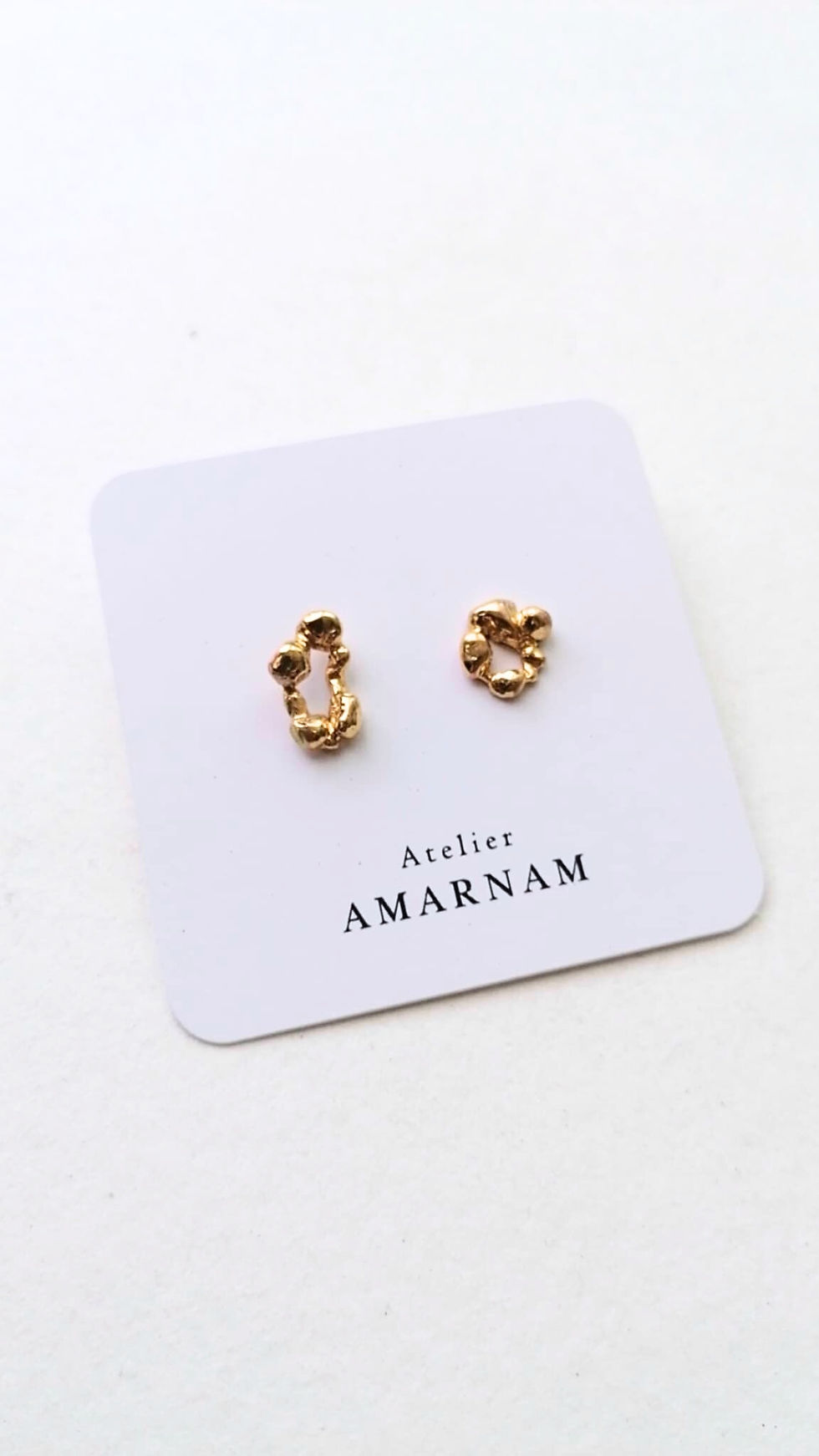 Gold earrings on a white card with "Atelier AMARNAM" text, set against a light background. Minimalist and elegant design.