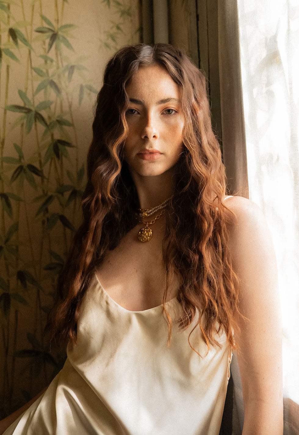 Woman with long wavy hair, wearing gold necklaces and a beige dress. She stands against a bamboo-patterned wall, looking serene.