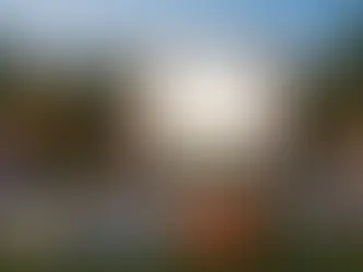 An outdoor backyard movie party scene featuring a large inflatable screen in the center of a grassy lawn. Several children are gathered on blankets, pillows, and bean bags facing the screen, while two adults sit in lounge chairs in the foreground. To the left is a colorful castle-style bounce house, and to the right is a table with a popcorn machine. Festive string lights with pennant flags and balloons decorate the yard, creating a cheerful evening atmosphere.