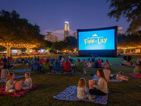 An outdoor movie screening in a city park during the evening, featuring a large inflatable screen displaying the title “The Adventures of Finn & Lily.” Families and friends sit on blankets and lawn chairs, surrounded by string lights wrapped around trees. Food trucks are visible in the background, and tall buildings glow in the distance, creating a festive, family-friendly atmosphere.
