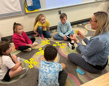 A teacher sits with five elementary students, doing an activity.