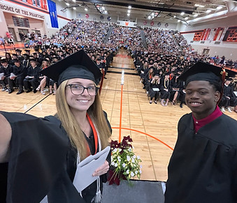 Two graduates taking a selfie in front of the crowd at the 2025 commencement ceremony in the gym.