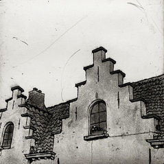 Looking up at two identical step-gabled tops of buildings in the winding streets of Brrugge. Each gable sports a window, curved at the top, and we can see a small window box with flowers in the closer of the two. The sky is bright but overcast. There is a mood about it.