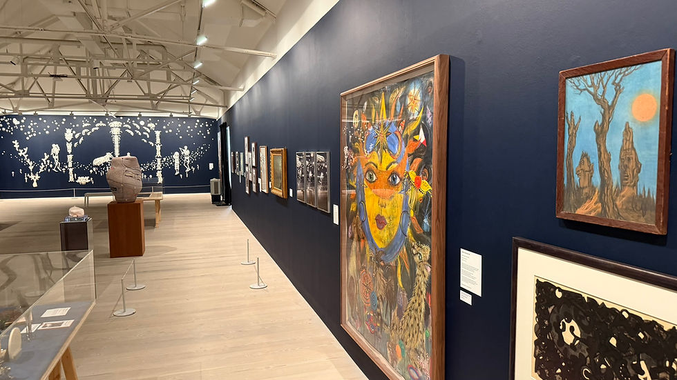 Art gallery with dark blue walls, framed paintings and sculptures, including a colorful face portrait and skeletal wall mural. Saatchi Gallery, The Sun and The Moon Exhibition, London, June, 2026. Aradia, speak to me' by artist Freya Pocklington, featured in the Saatchi Gallery exhibition "The Sun and The Moon: Art Inspired by the Celestial"