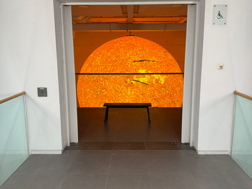 A hallway opens to a room with a glowing orange sun display, a bench in front, and a wheelchair-accessible sign on the wall. Luke Jerram, Helios, installed at Saatchi Gallery for The Sun and The Moon: Art Inspired by the Celestial, London, 2026.