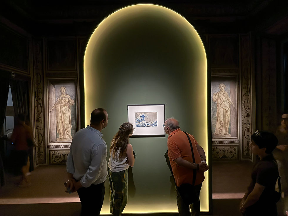 Museum visitors study a framed wave print in a dim gallery, flanked by classical wall paintings and a glowing arch. Katsushika Hokusai's 'The Great Wave Off Kanagawa, 1831, part of the Thirty-six Views of Mount Fuji series, as presented at Palazzo Bonaparte, Rome, June 2026. The Fine Art Ledger
