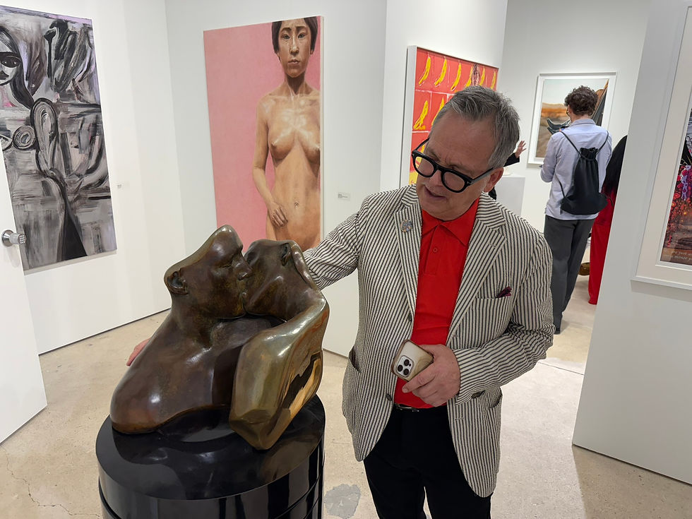 Man in striped suit admires sculpture of two faces kissing in an art gallery, with colorful paintings in the background. Ethan Cohen Gallery. Art Miami. Carole Feuerman Sculpture. The Fine Art Ledger
