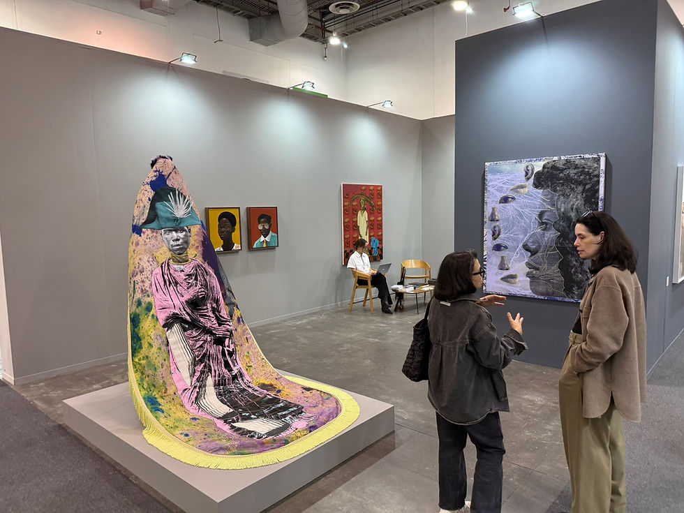 Art fair scene with two women discussing a large, colorful artwork. Paintings on walls; a seated person in the background.The name of this work by Raphaël Barontini's Même sur ces cendres, je vous combattrai encore (2025). Presented by The work was featured at the Zona Maco art fair in Mexico City by Mariane Ibrahim Gallery. The Fine Art Led ger. Artwork Passports.