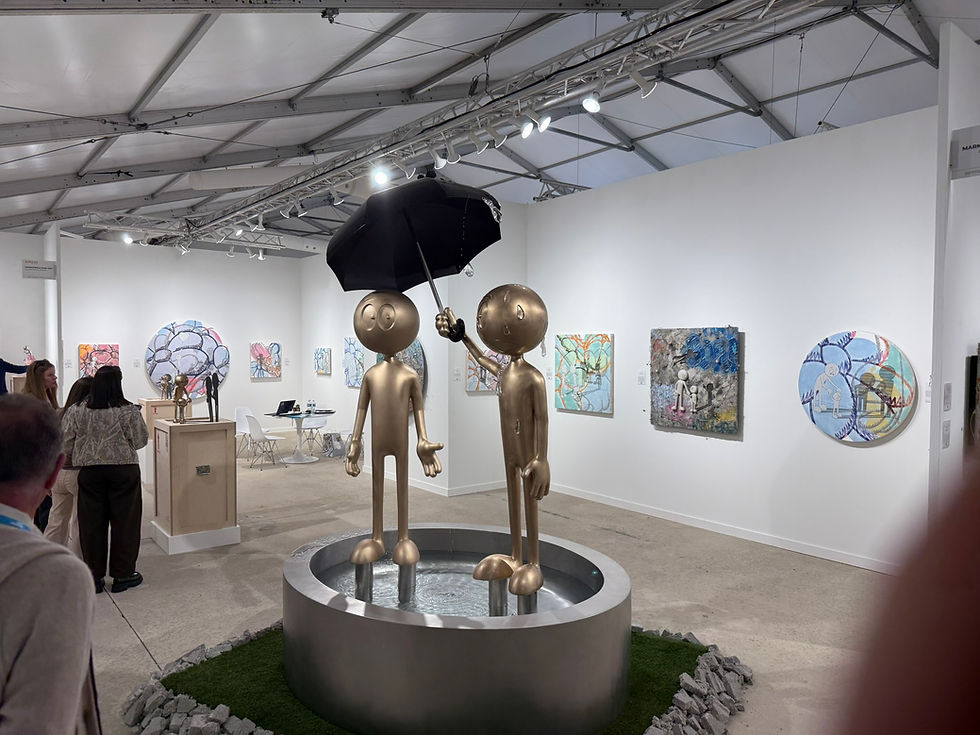 Two golden sculptures, one holding an umbrella, stand in a gallery. Colorful abstract paintings on white walls surround them.  'You Come First', Kai, Markowicz Fine Art, Art Miami, December 2025

