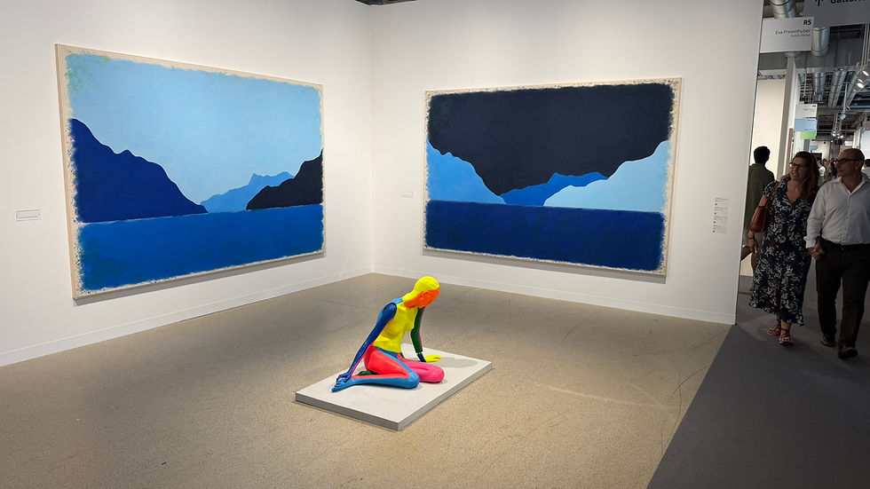 Art gallery with two large blue landscape paintings and a bright multicolor kneeling sculpture; visitors walk by. Ugo Rondinone, 'Nude (xxxxxxxxxxxxx) (rainbow)' , 2021 and 'Neuntermaizweitausendundeinundzwanzig', 2026. Galerie-Eva-Presenhuber. Art Basel Switzerland June 2026