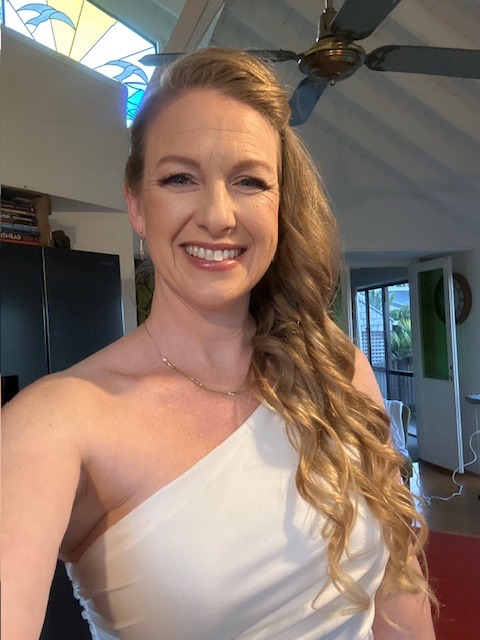 Smiling woman with long blonde curls and natural makeup in a one-shoulder white top indoors