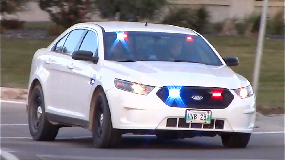 A Winnipeg white police car with red and blue lights on, driving on a road. Green grass blurred in the background.