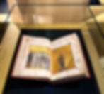 The illuminated manuscript of Panoplia Dogmatike with colorful religious figures and gold backgrounds displayed in a glass case on a wooden surface.
