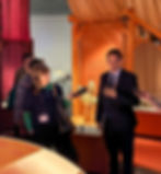 Peter Frankopan in a suit speaks to reporters holding microphones in an exhibit with orange lighting and sculptures. Text is visible in the background.