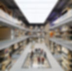 People stand on a glass floor in a multilevel gallery inside V&A East Storehouse filled with shelves of artifacts. Bright lighting and industrial design.