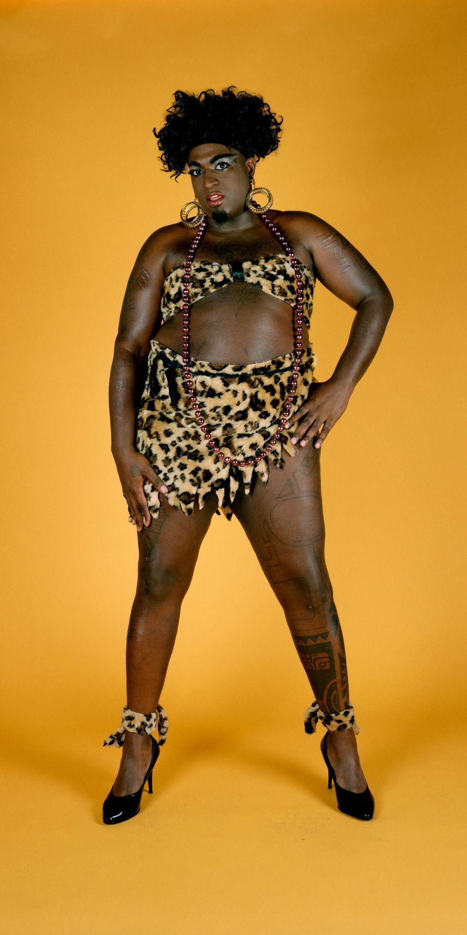 Person in leopard print outfit and heels poses confidently against an orange backdrop. They have curly hair, tattoos, and bold makeup.