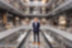 Tim Reeve in suit stands on glass floor in multi-level gallery with displayed artifacts. The setting is industrial, organized, with "Weston Collections Hall" visible.