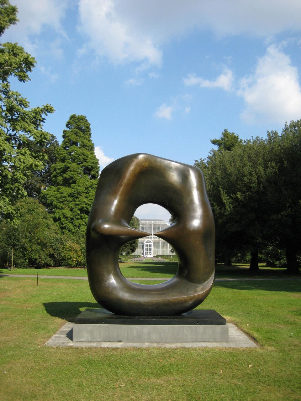 Abstract bronze sculpture by Henry Moore on a stone base in a green park. Trees line the background under a blue sky with clouds. No text visible.