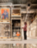 Person in a checkered sweater examines guitars in a modern storage room in V&A East Storehouse. Paintings and industrial pipes are visible, creating a curated environment.