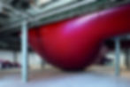 Large red inflated structure in industrial setting with concrete pillars and exposed pipes. Abstract art installation by Anish Kapoor in a spacious gallery.