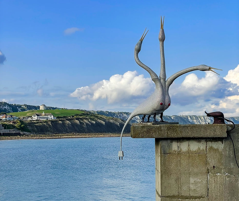 Strange gray bird-like sculpture by Laure Prouvost with red eyes on a pier over calm sea, with cliffs and blue sky in the background
