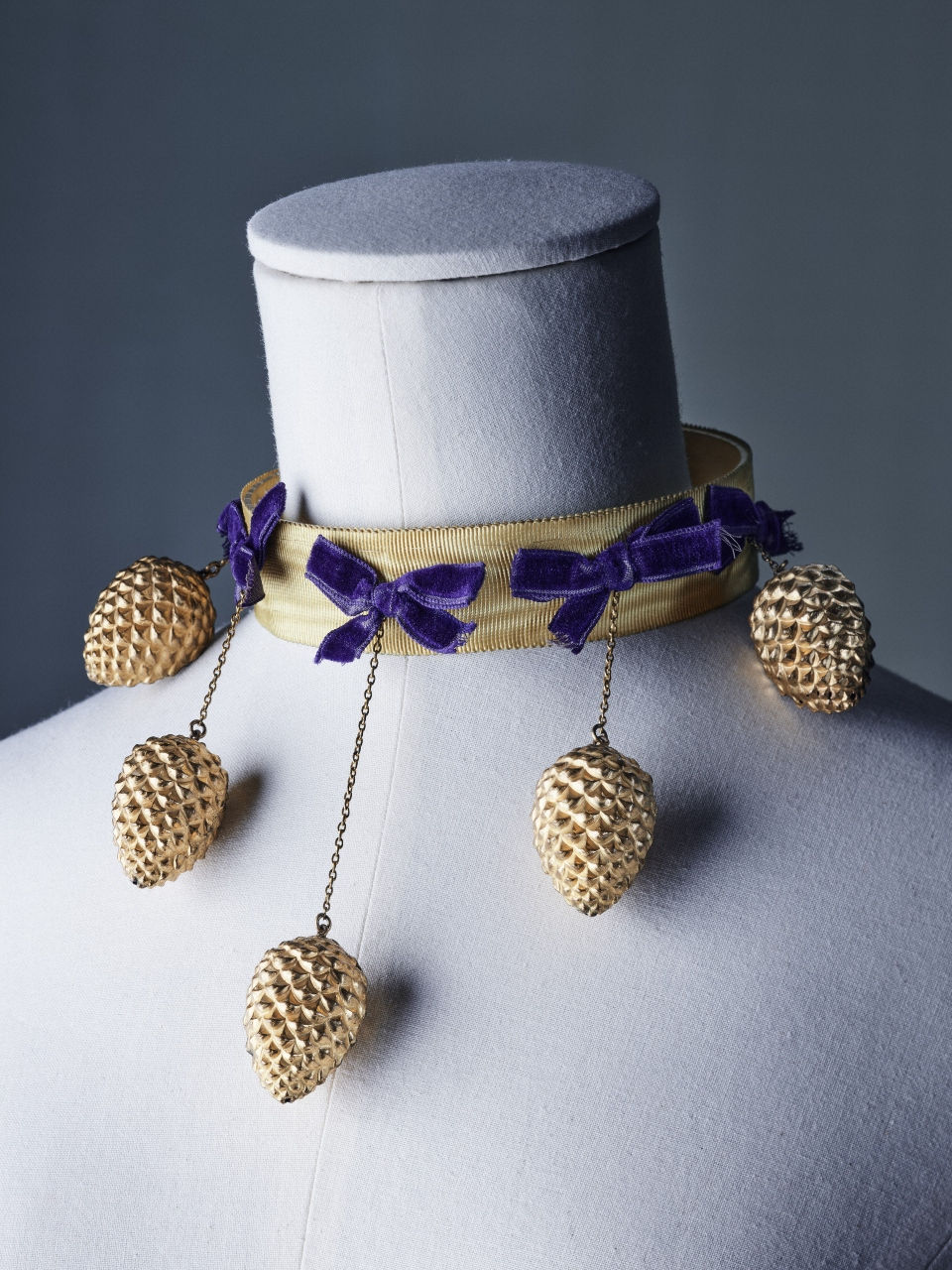 Mannequin adorned with a gold Schiaparelli choker featuring purple bows and hanging pinecone charms, set against a neutral gray background.