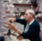 Henry Moore sculpting with clay in a cluttered studio. Focused expression. Shelves filled with various sculptures. Can labeled "Rawlplug Plastic Wood."