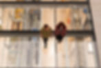 Two people walk on a glass floor in the V&A East Storehouse, viewing art below. One wears a red checkered jacket. Visible text: "AGRA COLONNADE."