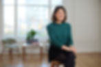 Smiling Alice Regent in a green sweater sits on a stool in a bright room with a large window, wooden floor, and chairs. A plant is on a table.