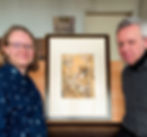 Two people smiling stand on either side of a framed artwork depicting a mythological scene. Books and a poster are in the background.