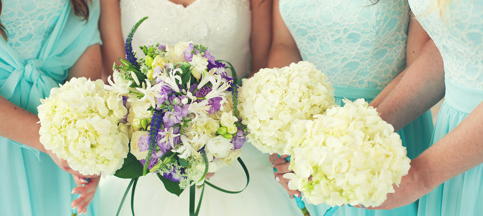 Bride holding a bouquet with soft purple accents, alongside bridesmaids carrying hand tied bouquets made exclusively with white hydrangeas.