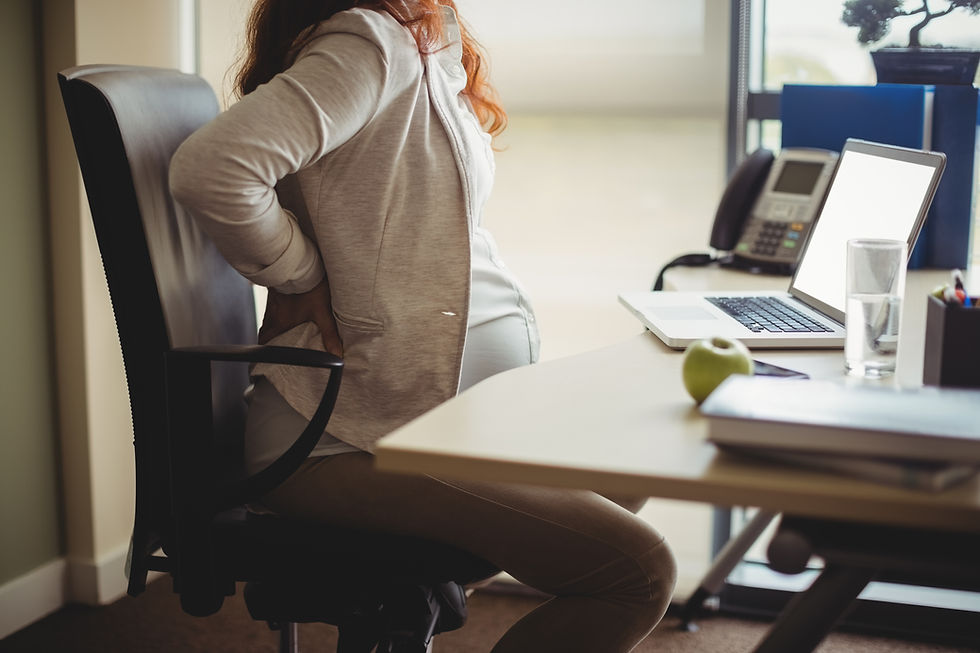 Mulher grávida sentada em uma cadeira com as mãos na costa. Ela está em uma mesa de escritório, com um notebook na sua frente