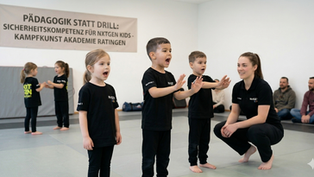 Eine Gruppe von drei jungen Kindern im NxtGEN Kids Panda-Training (Kampfkunst Akademie Ratingen) übt in einer Reihe auf grauen Matten, laut zu sprechen. Ein Junge in der Mitte schreit mit offenem Mund, flankiert von zwei weiteren Kindern, während eine Trainerin (Crouching slightly) sie mit positiver Verstärkung und spezifischem Feedback ermutigt. Ein großes Banner an der Wand im Hintergrund nennt die Prinzipien des Trainings: 'PÄDAGOGIK STATT DRILL: SICHERHEITSKOMPETENZ FÜR NXTGEN KIDS - KAMPFKUNST AKADEMIE RATINGEN'. Im Hintergrund sind weitere Kinder und Eltern zu sehen.