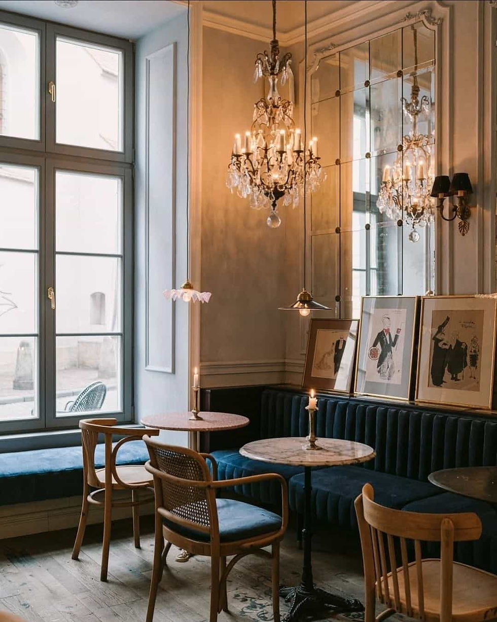 Elegant café interior with wooden chairs, round marble tables, lit candles, chandeliers, framed art, and large windows. Cozy and vintage atmosphere.