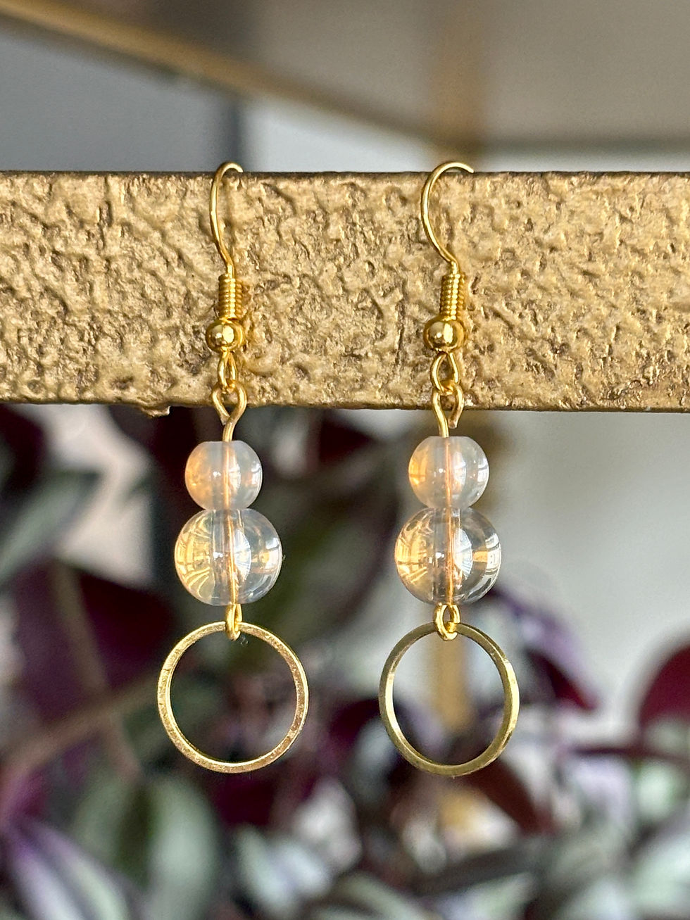 Thumbnail: Hanging from a gold bar with a plant in the background is a pair of dangle earrings with two champagne beads stacked on top of each other with a gold metal circle hanging at the bottom. The sizing of the beads and the metal circle go from smallest to largest, almost like a snowman.