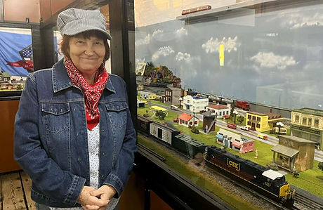 Carolyn McKinney as "Conductor Clara" in railroader's hat, standing by model train display.