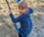 Girl on a blue swing in a sandy playground, wearing a dark shirt with colorful butterflies, looking back with a curious expression.